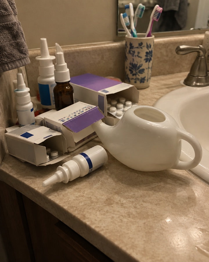 Bathroom counter crowded with nasal sprays, pills, and a neti pot